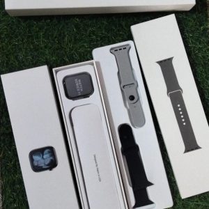 Premium Apple Watch Series 11 unboxed on grass showing the main dial with Apple logo branding box, dual silicone straps, and wireless charger