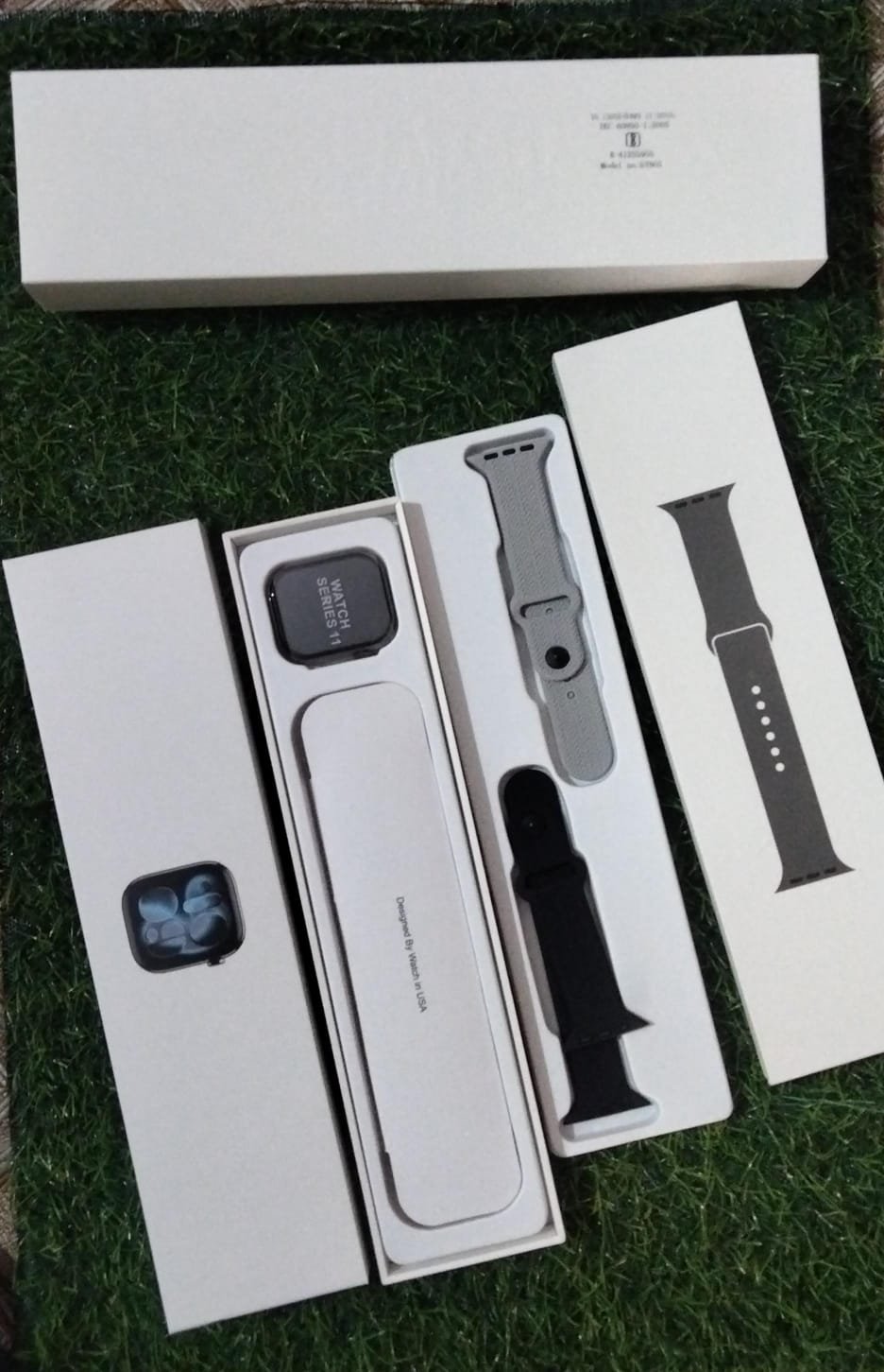 Premium Apple Watch Series 11 unboxed on grass showing the main dial with Apple logo branding box, dual silicone straps, and wireless charger